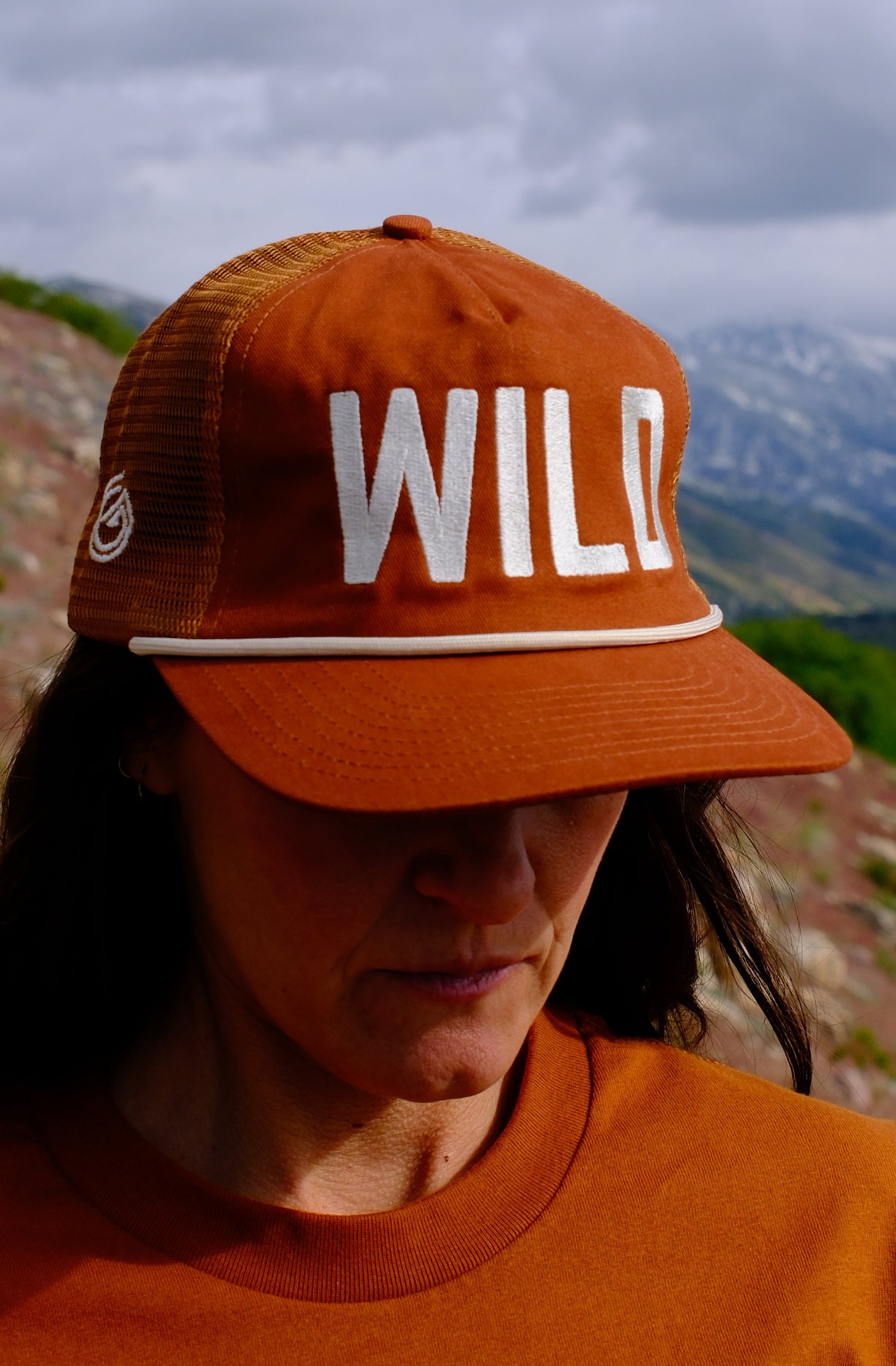 Person wearing a brown cap and shirt with a logo, standing in a mountainous landscape.