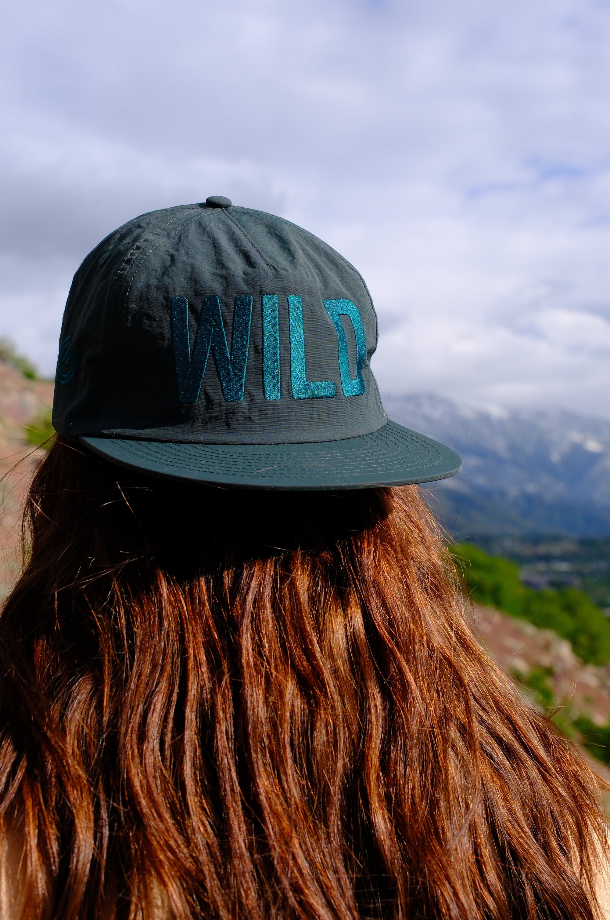 Person wearing a cap with 'WILD' on it, standing against a mountainous landscape.